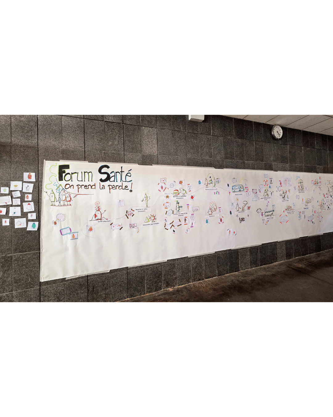 Une fresque de plusieurs mètres est accrochée sur un mur. Le titre est "Forum santé". Il y a beaucoup de dessins accrochés et dessinés sur la fresque.