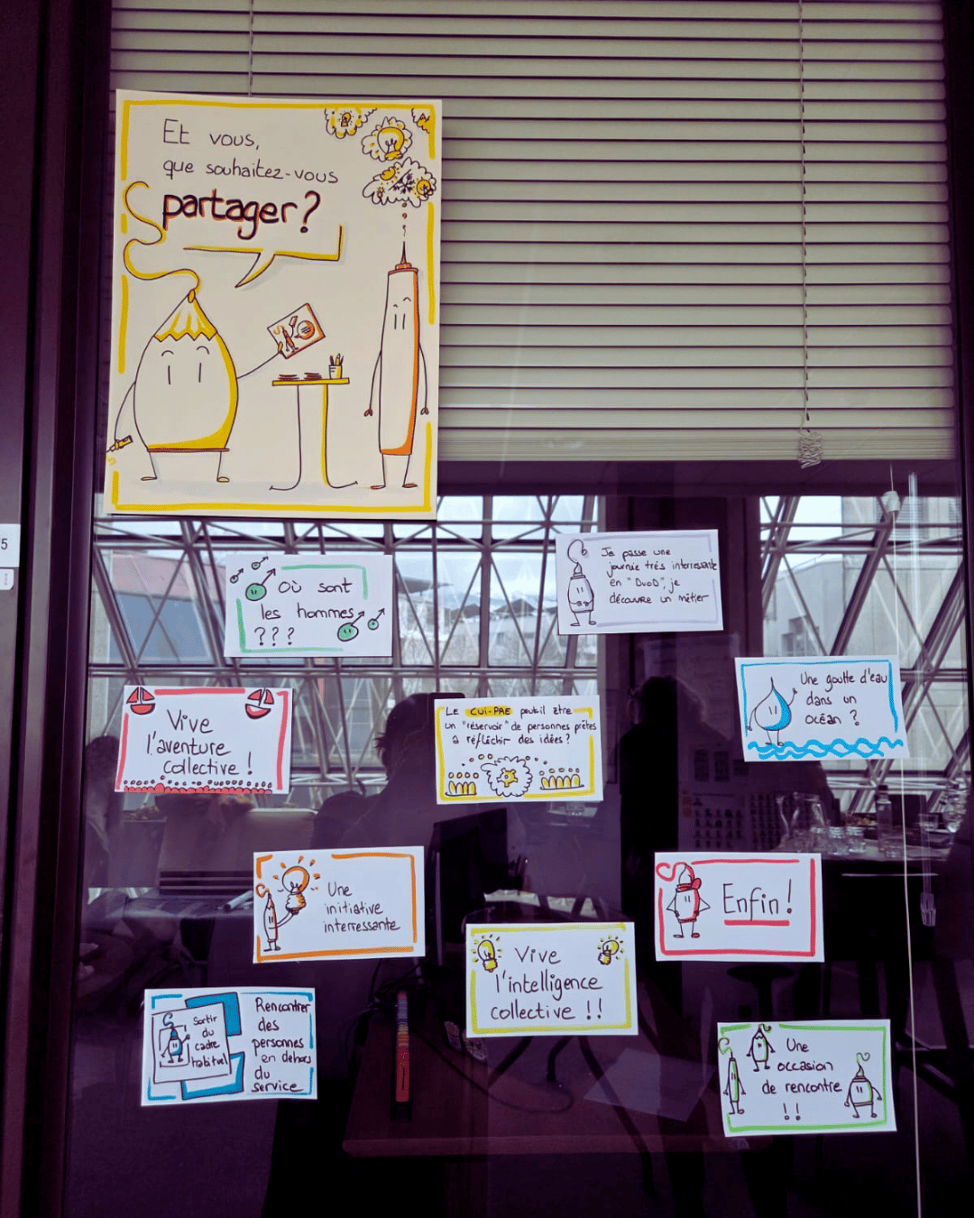 Une vitre sur laquelle sont accrochés une affiche et des petites cartes. Sur l'affiche on peut voir deux personnages crayons, dont l'un tiens une petite carte et dit "Et vous, que souhaitez-vous partager ?" Sur les cartes, on peut voir les participations des personnes. Elles sont très colorées.