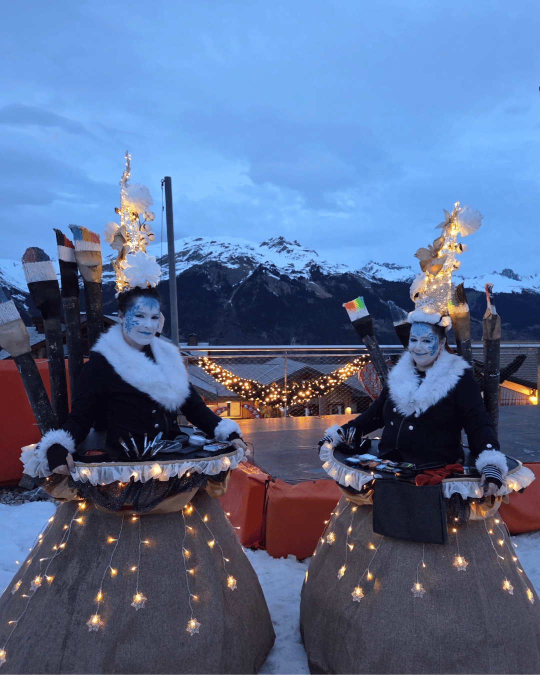 Deux maquilleuses en costumes d'hiver nous font face. Il fait presque nuit et on voit les montagnes enneigées en arrière plan. Elles ont de très hautes coiffes lumineuses et des grandes robes à crinoline, elles aussi illuminées
