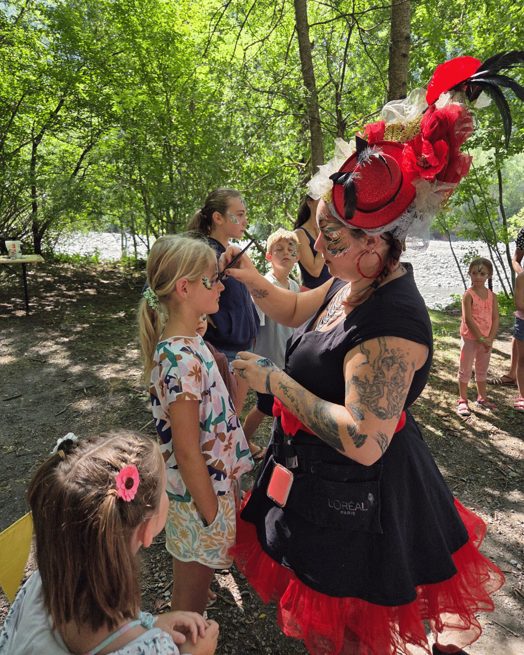 Une maquilleuse maquille des personnes qui font une ronde autour d'elle. Elle est debout, a une coiffe faite de plumes et de fleurs et une robe rockabilly. Le tout est rouge et noir.