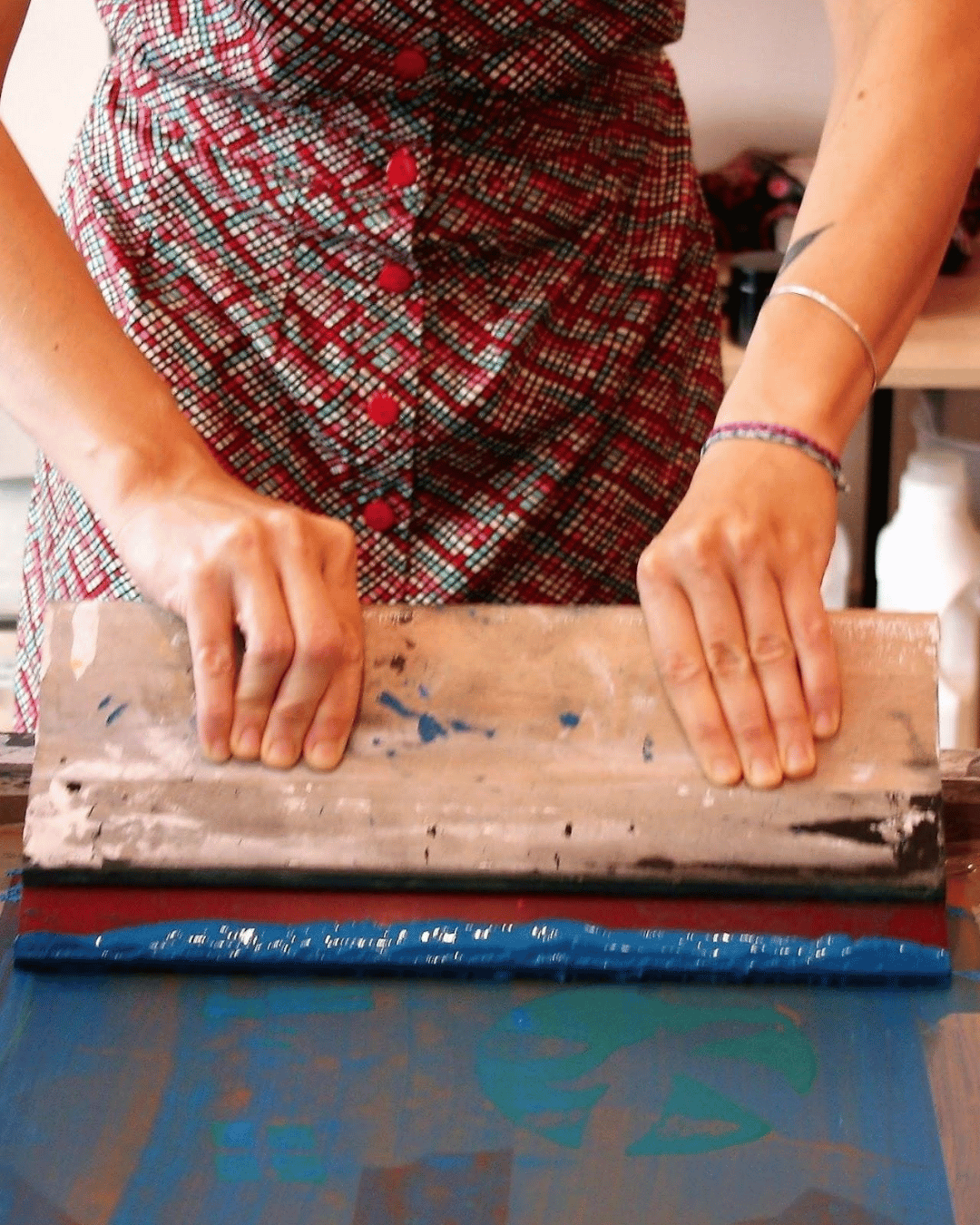 Une personne avec un vêtement coloré est en train de racler un cadre de sérigraphie avec de l'encre bleue, en utilisant une grosse raclette en bois. On ne vois que les bras et une partie du corps de la personne