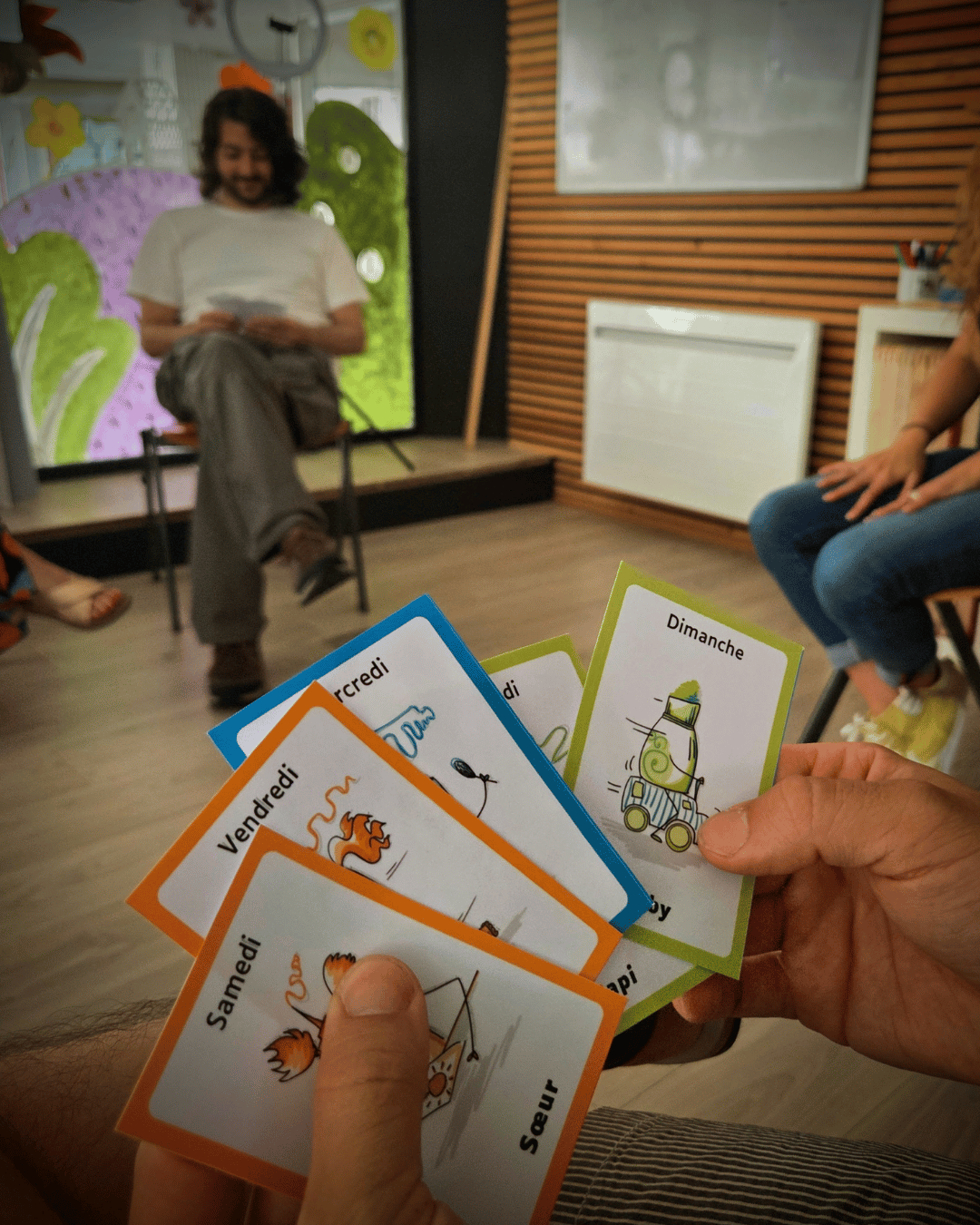 Au premier plan, on voit un jeu de carte avec des personnages aux formes de crayons. C'est un jeu de 7 familles.